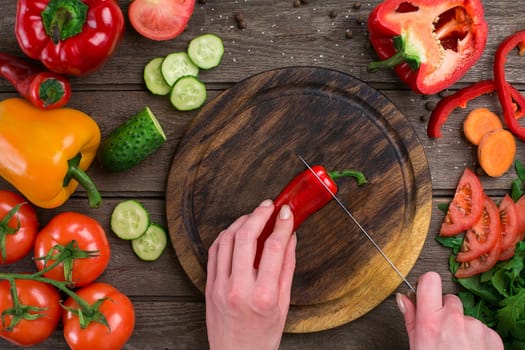Female hands cutting hot peppers at table, top view. On the table vegetables and a wooden board