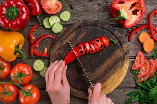 Female hands cutting hot peppers at table, top view. On the table vegetables and a wooden board