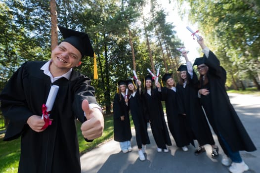 Happy young caucasian male graduate showing thumbs up. A group of graduate students outdoors