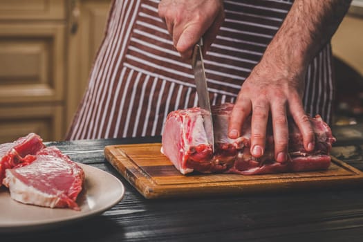 Man cuts of fresh piece of meat on a wooden cutting board in the home kitchen. A man in a striped apron with a big knife in his hands