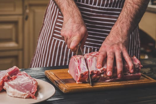 Man cuts of fresh piece of meat on a wooden cutting board in the home kitchen. A man in a striped apron with a big knife in his hands