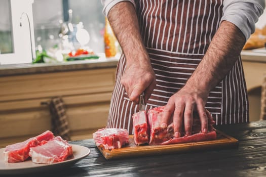 Man cuts of fresh piece of beef on a wooden cutting board in the home kitchen. A man in a striped apron with a big knife in his hands