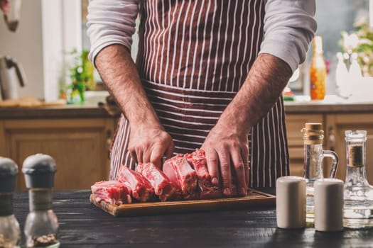 Man cuts of fresh piece of meat on a wooden cutting board in the home kitchen. A man in a striped apron with a big knife in his hands