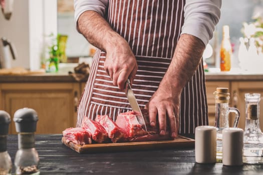 Man cuts of fresh piece of meat on a wooden cutting board in the home kitchen. A man in a striped apron with a big knife in his hands