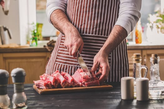 Man cuts of fresh piece of beef on a wooden cutting board in the home kitchen. A man in a striped apron with a big knife in his hands