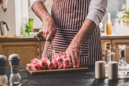 Man cuts of fresh piece of meat on a wooden cutting board in the home kitchen. A man in a striped apron with a big knife in his hands