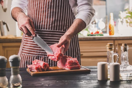 Man cuts of fresh piece of meat on a wooden cutting board in the home kitchen. A man in a striped apron with a big knife in his hands