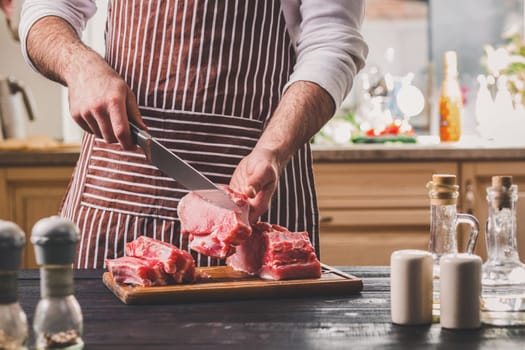 Man cuts of fresh piece of meat on a wooden cutting board in the home kitchen. A man in a striped apron with a big knife in his hands