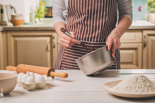 Young woman holding bowl with dough and whisk, closeup. A woman in a striped apron is cooking in the kitchen