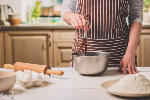 Young woman holding bowl with dough and whisk, closeup. A woman in a striped apron is cooking in the kitchen
