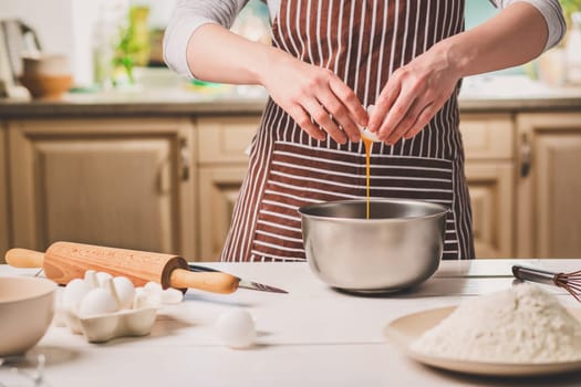 Young woman breaking egg over bowl with dough, close-up. A woman in a striped apron is cooking in the kitchen