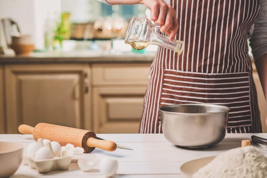 Young woman pouring oil into a bowl with dough, close-up. A woman in a striped apron is cooking in the kitchen