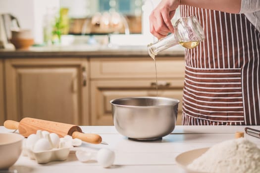 Young woman pouring oil into a bowl with dough, close-up. A woman in a striped apron is cooking in the kitchen