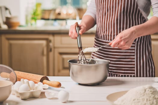 Young woman holding bowl with dough and whisk, closeup. A woman in a striped apron is cooking in the kitchen