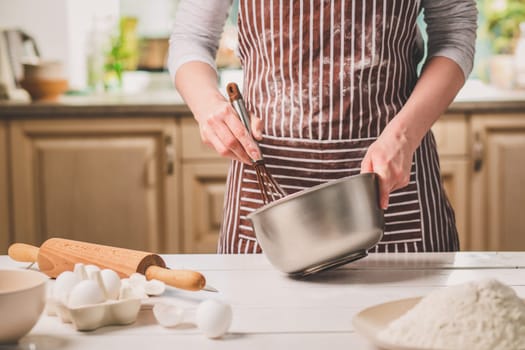 Young woman holding bowl with dough and whisk, closeup. A woman in a striped apron is cooking in the kitchen