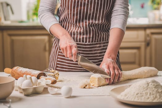 Close-up view of two woman's hands cut knife dough. A woman in a striped apron is cooking in the kitchen