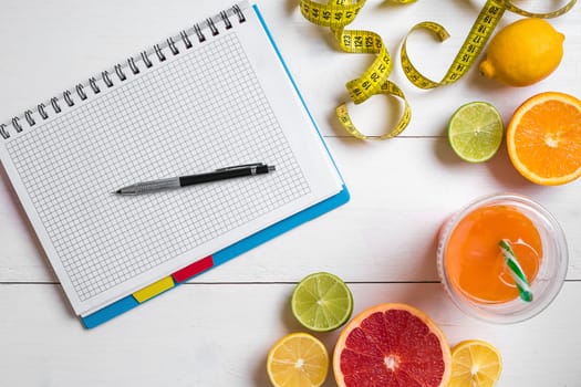 Fresh juice in glass from citrus fruits - lemon, grapefruit, orange, notebook with pencil on white wooden background, top view