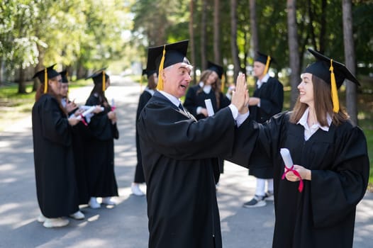 A group of graduates in robes outdoors. An elderly man and a young woman congratulate each other on their graduation