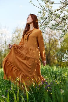 a happy, slender, sweet woman stands in a long orange dress in the tall grass near a flowering tree and happily smiling lifts the hem of her dress. High quality photo