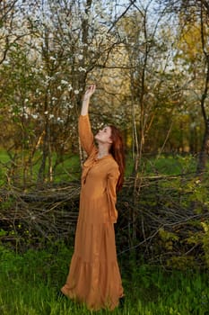 an elegant, sophisticated woman poses standing near a wicker fence in a dacha in a long orange dress, raising her hands up. High quality photo