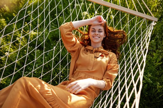 a beautiful, elegant woman lies in a long orange dress on a mesh hammock resting in nature, illuminated by the warm sunset light, smiling happily holding her hand near her forehead. High quality photo