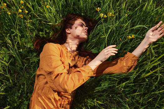 a calm woman with long red hair lies in a green field of tall grass in an orange dress with her eyes closed, stretching her arms in front of her. High quality photo