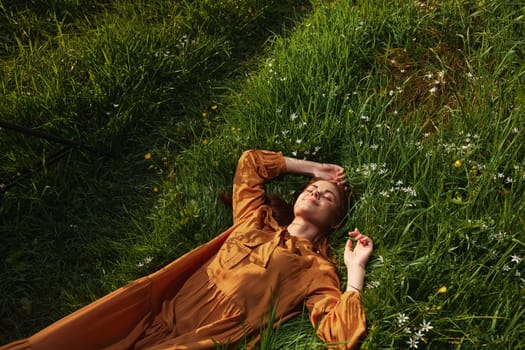 a happy, relaxed woman, resting lying in the green grass, in a long orange dress, with her eyes closed and a pleasant smile on her face, holding her hands near her face, enjoying harmony with nature and recuperating. High quality photo