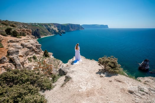 Woman in a white dress on the sea. Side view Young beautiful sensual woman in white long dress posing on a rock high above the sea at sunset. Girl in nature against the blue sky.