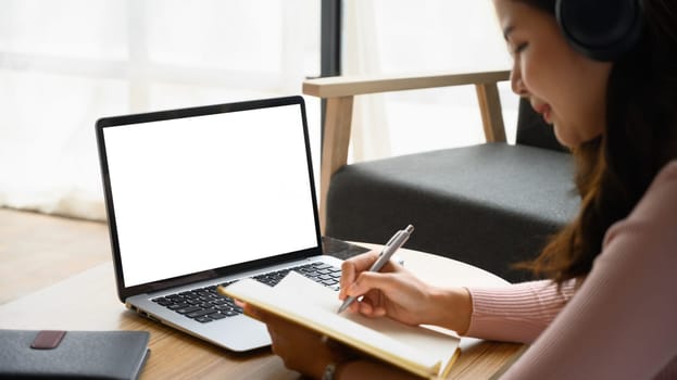 Concentrated young woman having distance learning online virtual class on laptop and making notes on notebook.