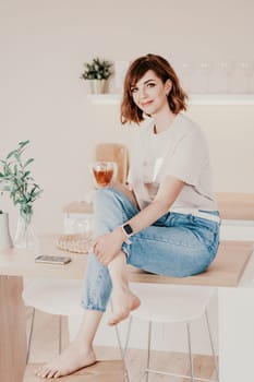 Woman kitchen tea. Carefree woman rests in a cozy kitchen, drinks tea, sits on the table and dreams, free space. Woman holding cup while enjoying hot drink at home.