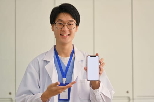 Smiling male doctor in uniform holding smartphone screen, presenting blank smartphone display for medical advertisement, mock up.