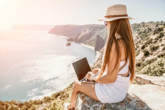 Successful business woman in yellow hat working on laptop by the sea. Pretty lady typing on computer at summer day outdoors. Freelance, travel and holidays concept.