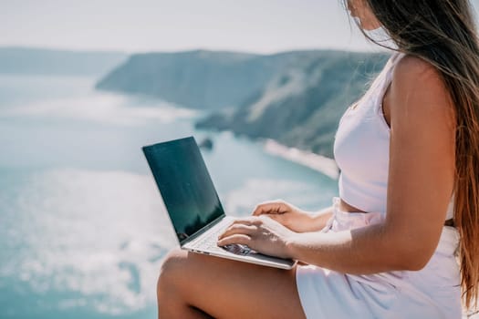 Successful business woman in yellow hat working on laptop by the sea. Pretty lady typing on computer at summer day outdoors. Freelance, travel and holidays concept.