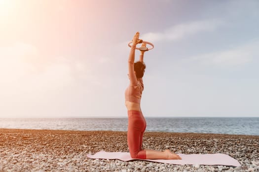 Middle aged well looking woman with black hair doing Pilates with the ring on the yoga mat near the sea on the pebble beach. Female fitness yoga concept. Healthy lifestyle, harmony and meditation.