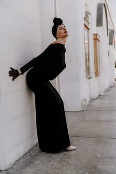 Stylish woman in the city. Fashion photo of a beautiful model in an elegant black dress posing against the backdrop of a building on a city street.