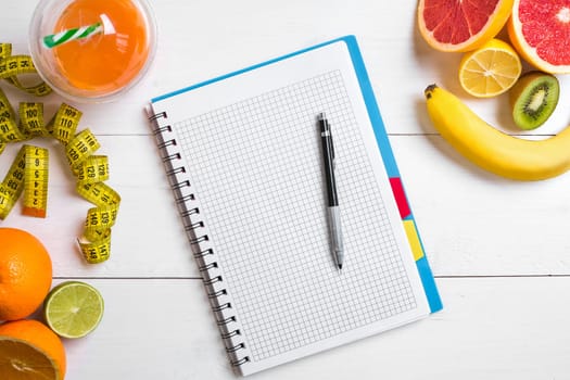 Fresh juice in glass from citrus fruits - lemon, grapefruit, orange, notebook with pencil on white wooden background, top view