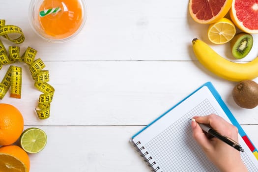 Fresh juice in glass from citrus fruits - lemon, grapefruit, orange, notebook with pencil on white wooden background, top view