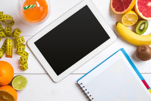 Fresh juice in glass from citrus fruits - lemon, grapefruit, orange, notebook with pencil on white wooden background, top view. White tablet with copy space