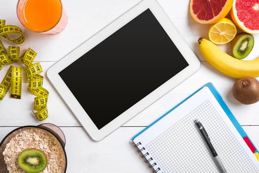 Fresh juice in glass from citrus fruits - lemon, grapefruit, orange, notebook with pencil on white wooden background, top view. White tablet with copy space