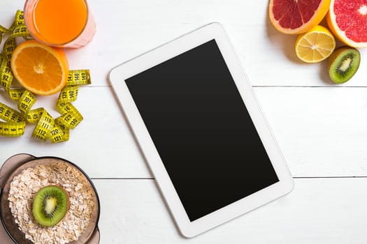 Fresh juice in glass from citrus fruits - lemon, grapefruit, orange, bowl of oatmeal on white wooden background, top view. White tablet with copy space