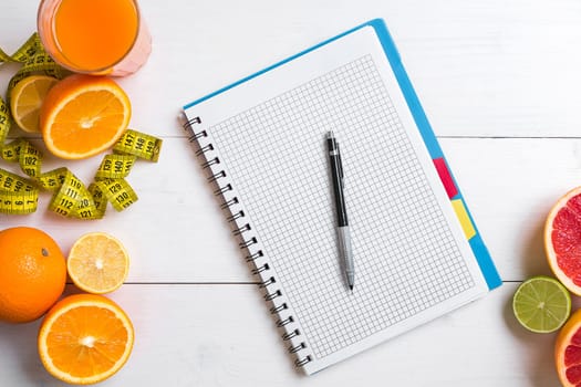 Fresh juice in glass from citrus fruits - lemon, grapefruit, orange, notebook with pencil on white wooden background, top view