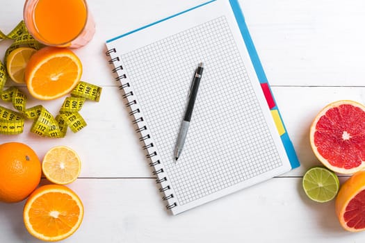 Fresh juice in glass from citrus fruits - lemon, grapefruit, orange, notebook with pencil on white wooden background, top view