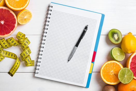 Fresh juice in glass from citrus fruits - lemon, grapefruit, orange, notebook with pencil on white wooden background, top view