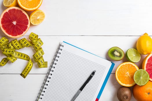 Fresh juice in glass from citrus fruits - lemon, grapefruit, orange, notebook with pencil on white wooden background, top view