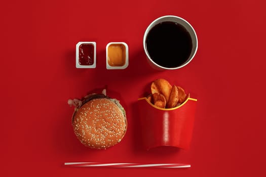 Fast food dish top view. Meat burger, potato chips and glass of drink on red background. Takeaway composition. Wrapped French fries, hamburger, mayonnaise and ketchup sauces on red desk. Copy space