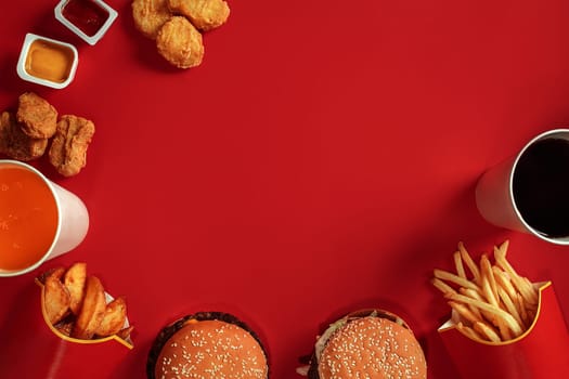 Fast food dish top view. Meat burger, potato chips and glass of drink on red background. Takeaway composition. Wrapped French fries, hamburger, mayonnaise and ketchup sauces on red desk. Copy space