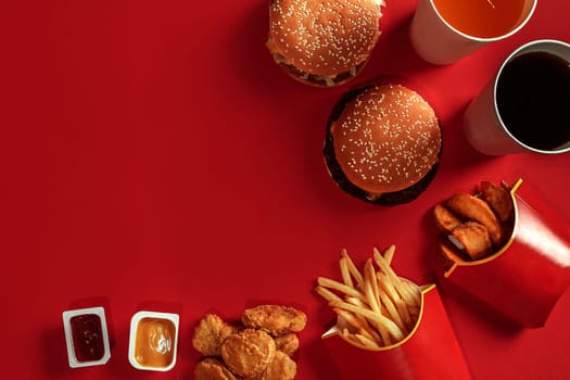 Fast food dish top view. Meat burger, potato chips and glass of drink on red background. Takeaway composition. Wrapped French fries, hamburger, mayonnaise and ketchup sauces on red desk. Copy space