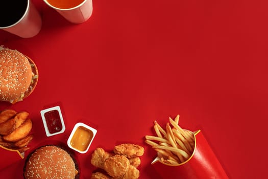 Fast food dish top view. Meat burger, potato chips and glass of drink on red background. Takeaway composition. Wrapped French fries, hamburger, mayonnaise and ketchup sauces on red desk. Copy space