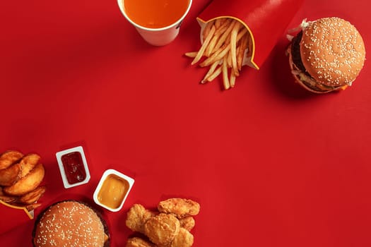 Fast food dish top view. Meat burger, potato chips and glass of drink on red background. Takeaway composition. Wrapped French fries, hamburger, mayonnaise and ketchup sauces on red desk. Copy space