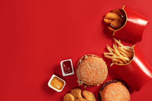 Fast food dish top view. Meat burger, potato chips and nuggets on red background. Takeaway composition. Wrapped French fries, hamburger, mayonnaise and ketchup sauces on red desk. Copy space